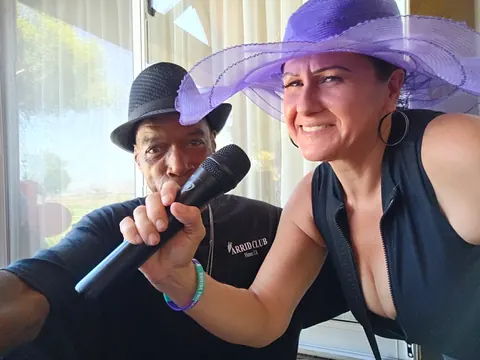 Two guests posing with a microphone, one wearing a wide purple hat.