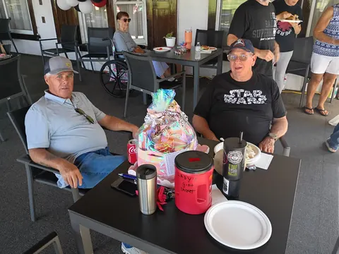 Two seated guests beside a wrapped gift basket and red raffle container.